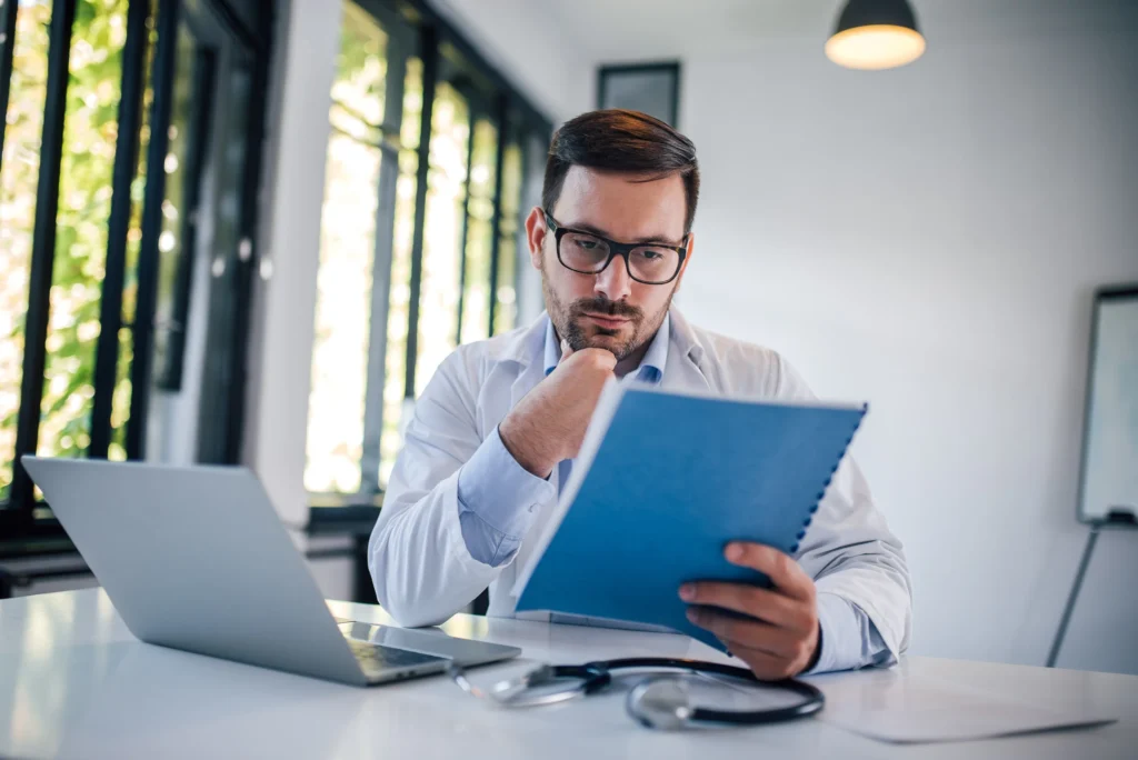A doctor looking at files with a computer in front of him.