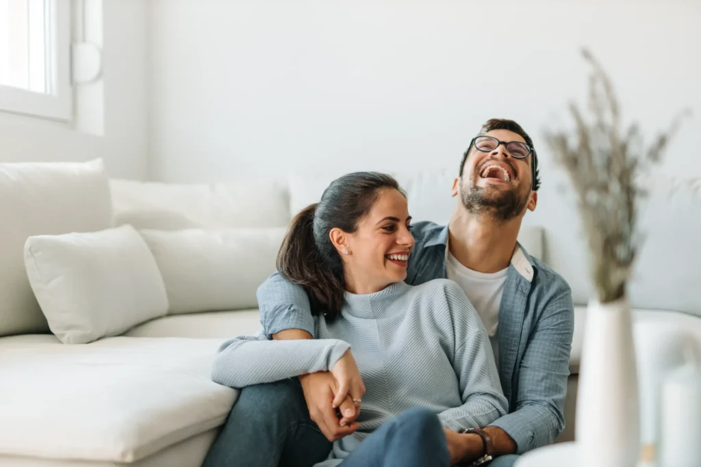 Happy couple sitting together and laughing in a bright living room, representing improved quality of life for those who benefit from kyphoplasty.