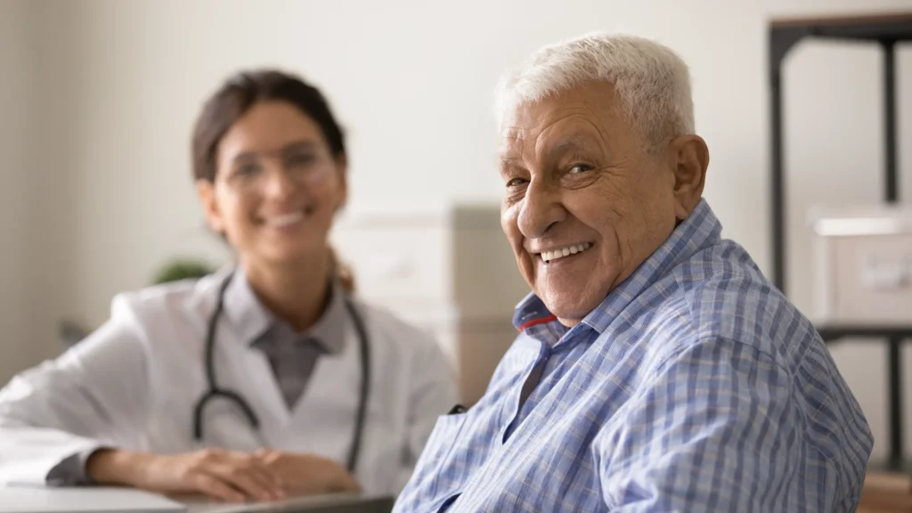 Smiling older man sitting in a medical office with a doctor in the background, representing discussions on how long relief lasts after RFA.